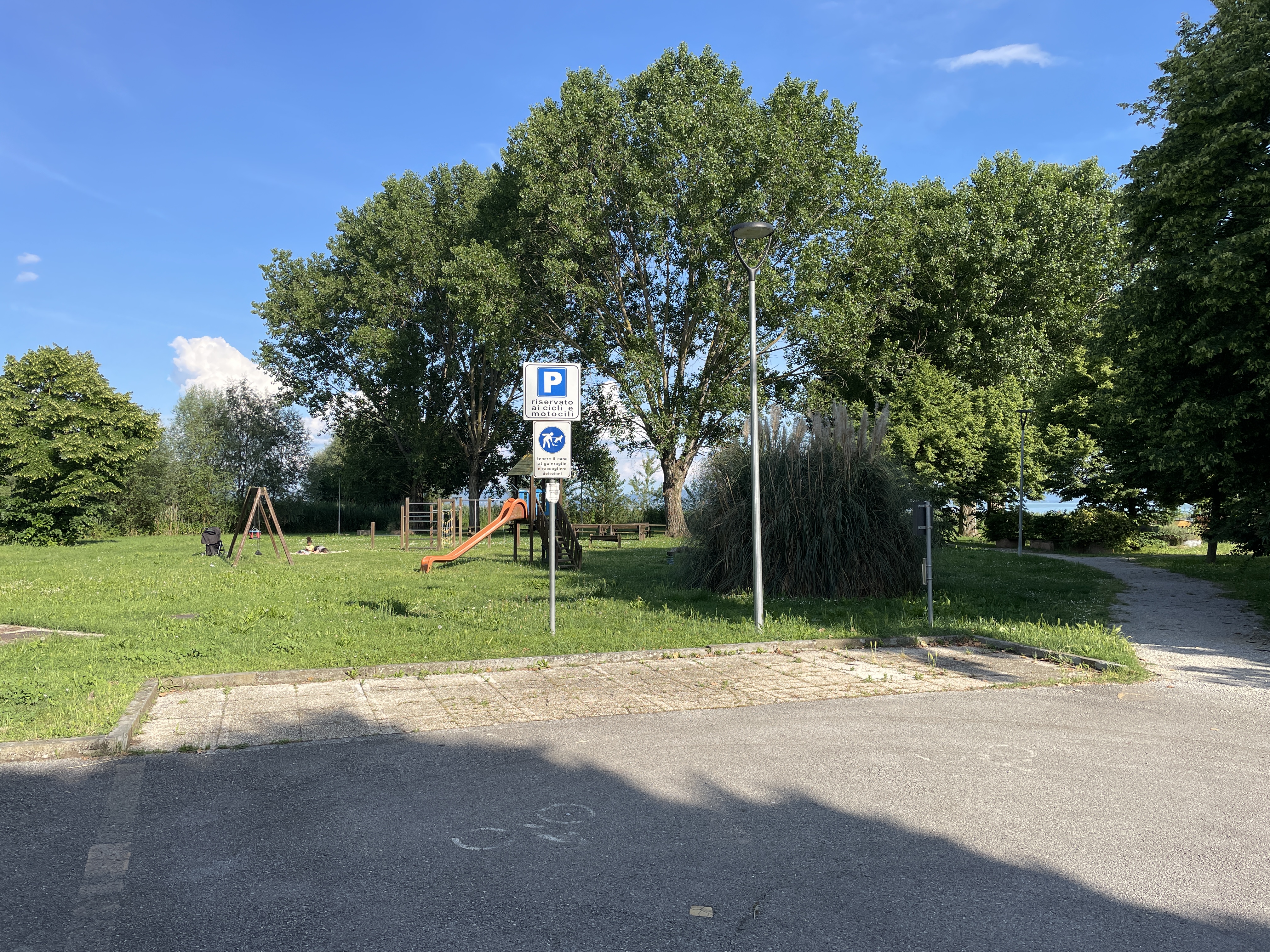 Parking de Borghetto avec des places réservées aux vélos et aux motos à côté d'un parc ombragé avec une aire de jeux. Autour, des arbres et des buissons.